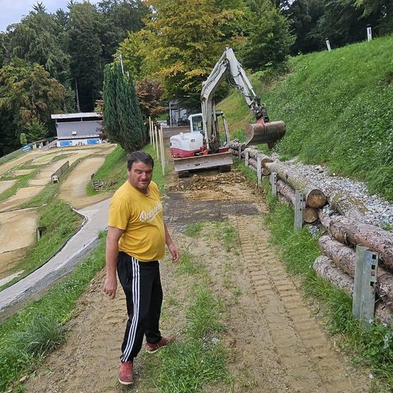 Ein Mann in einem gelben T-Shirt und schwarzen Hosen steht auf einem Lehmweg, mit einem Baufahrzeug neben ihm. Der Weg ist von Gras und Baumstämmen umgeben. Bäume und ein Gebäude sind im Hintergrund.