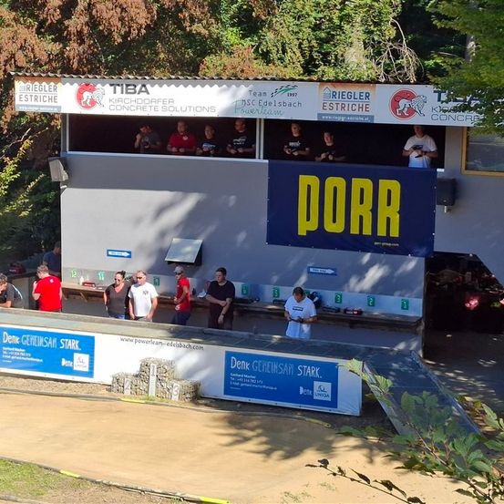 Eine Gruppe von Menschen steht vor einem Gebäude mit einem Banner, auf dem PORR steht. Es gibt Leute in der Kabine. Es gibt Bäume und Pflanzen in der Umgebung.