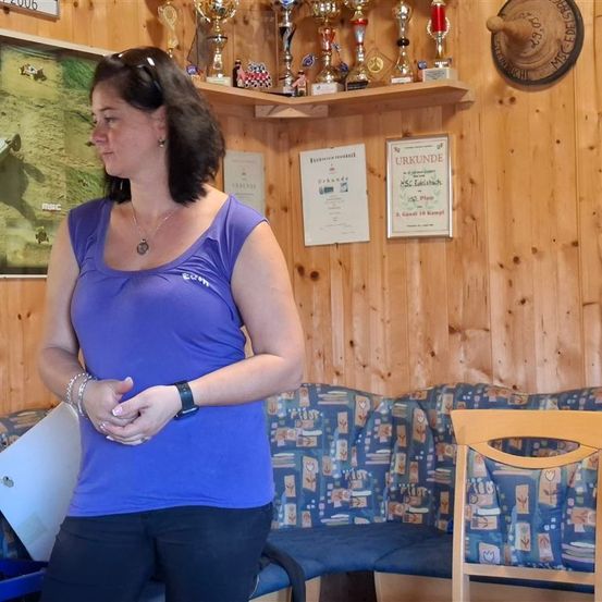 Eine Frau in einem lila Shirt steht in einem Raum mit Holzwänden. Sie trägt eine Halskette und Armbänder. Hinter ihr stehen Trophäen auf einem Regal. Ein gerahmtes Zertifikat hängt an der Wand.