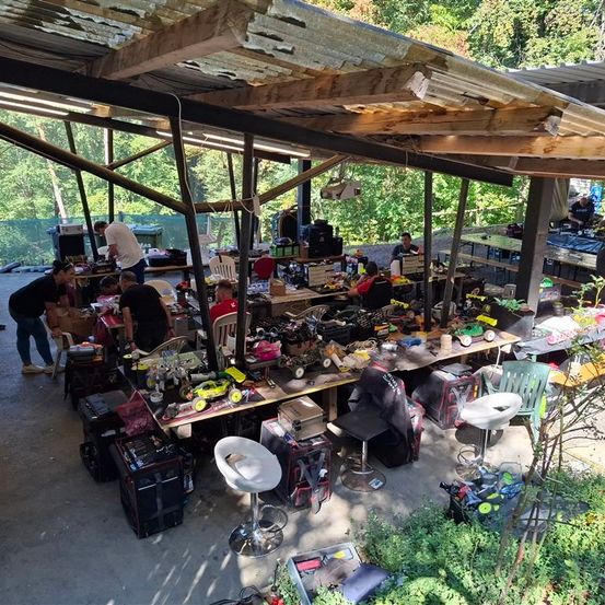 Ein Werkstattbereich mit mehreren Personen, die an verschiedenen Projekten arbeiten, unter einem hölzernen Vordach. Werkzeuge, Ausrüstung und verschiedene Gegenstände sind über mehrere Tische verteilt. Bäume und ein Zaun sind im Hintergrund sichtbar.