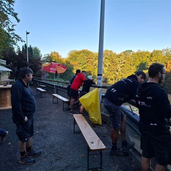 Eine Gruppe von Männern arbeitet an einer Struktur in der Nähe einer Bank und eines Tisches, mit Bäumen und einem blauen Himmel im Hintergrund. Ein Mann trägt ein rotes Hemd.