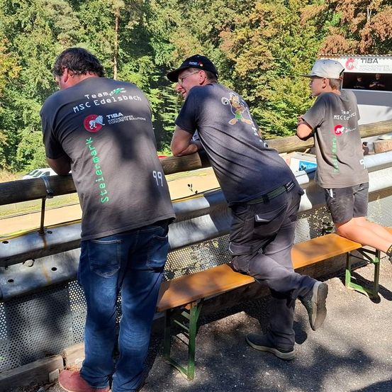 Drei Personen betrachten die Aussicht von einer Holzbarriere aus. Sie tragen T-Shirts mit verschiedenen Logos. Hinter ihnen steht ein LKW.