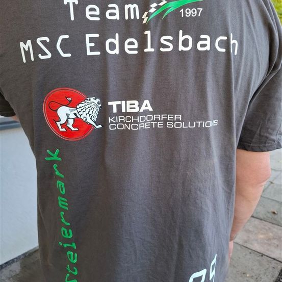 Ein graues Shirt mit dem Text Team 1997 MSC EDELSBACH. Es gibt ein Logo eines Löwen mit dem Text TIBA KIRCHDORFER CONCRETE SOLUTIONS. Auf der linken Seite steht der grüne Text STEIERMARK.