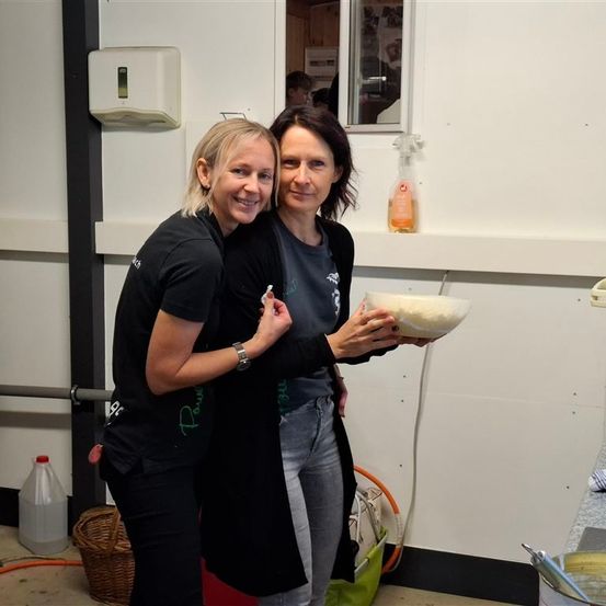 Zwei Frauen stehen zusammen, eine hält eine Schüssel mit Essen und lächelt. Sie sind in einem Küchenbereich mit einem Spiegel, einem Korb und Reinigungsmitteln.