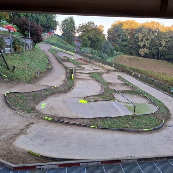 Luftaufnahme einer Motocross-Strecke mit einer kurvenreichen Strecke. Die Strecke ist von Gras und Bäumen umgeben, mit einem roten Baldachin in der Ferne.