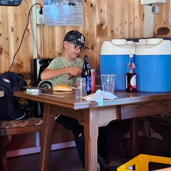 Ein junger Junge mit Brille sitzt an einem Holztisch und isst ein Sandwich. Flaschen, Dosen und Gläser stehen auf dem Tisch. Hinter ihm befinden sich zwei blau-weiße Kühlboxen. Ein Rucksack liegt auf einer Bank.
