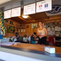 Eine Frau steht in einem kleinen Café und lächelt. Hinter ihr öffnet ein Junge eine Kiste, während ein anderer Junge sitzt. Ein VW-Logo ist an der Decke. Speisekarten und Poster schmücken die Wände.
