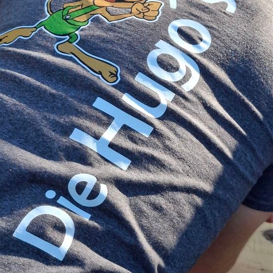 Ein graues T-Shirt mit 'Die Hugos' in weißer Schrift. Der Text wird von einer Cartoon-Zeichnung begleitet, möglicherweise ein Hund, in Grün und Braun. Der Schatten der Person ist sichtbar.