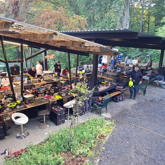 Ein Außenwerkstatt mit mehreren arbeitenden Personen, umgeben von mit Werkzeug und Ausrüstung gefüllten Tischen, unter einem Metallvordach.