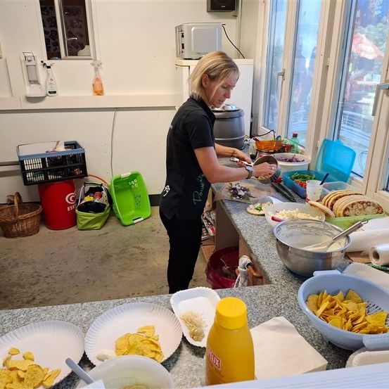 Eine Frau in einem schwarzen Shirt steht vor einem Tisch und bereitet Essen zu. Der Tisch ist mit verschiedenen Gerichten, Schüsseln und Tellern beladen. Hinter ihr befinden sich eine Mikrowelle, eine Aufbewahrungsbox und ein Korb. Die Szene spielt sich in einem Raum mit einem Fenster ab, das einen Blick nach draußen bietet.