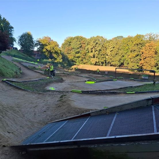 Eine Vogelperspektive auf eine Dirt-Strecke mit grünen Markierungen. Arbeiter in gelben Westen richten die Strecke ein. Die Strecke ist von Bäumen und Gras umgeben.