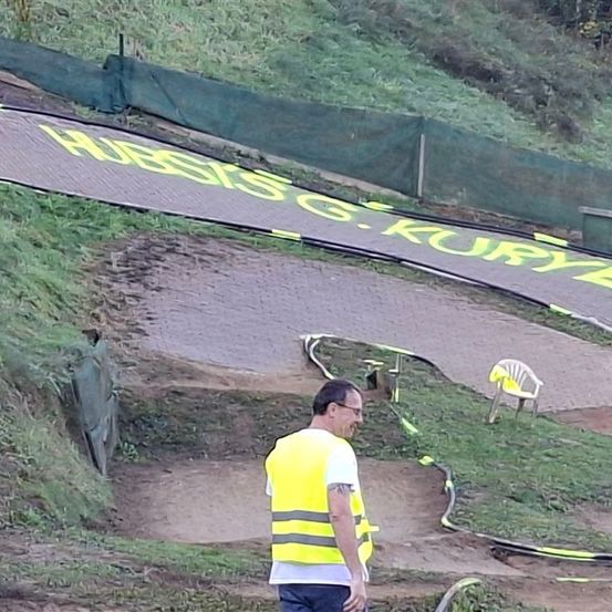 Ein Mann in einer gelben Weste steht auf einem schlammigen Pfad mit gelben Graffiti auf der Straße. Eines Stuhls ist auf der Seite platziert. Der Pfad ist mit grünen Netzen eingezäunt.