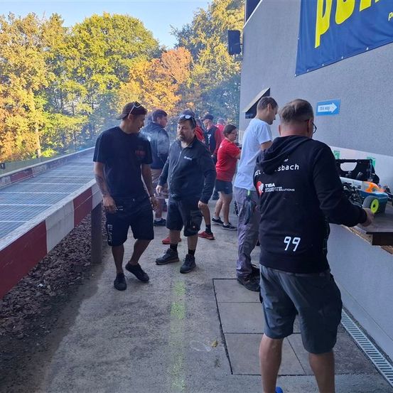 Eine Gruppe von Menschen ist draußen versammelt, einige tragen Sonnenbrillen. Ein Banner mit gelbem Text ist sichtbar. Sie stehen auf Beton mit einer Metallschiene auf der linken Seite.