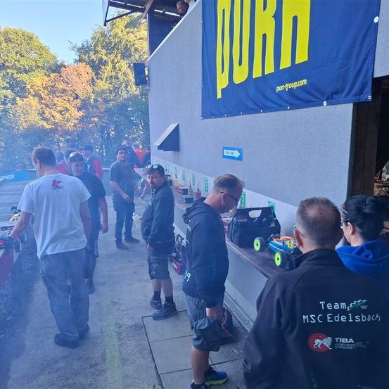 Eine Gruppe von Menschen steht vor einem Gebäude mit einem Banner, das "Purn" liest. Einige tragen Jacken, während andere Spielzeugautos tragen. In der Nähe steht eine Person in der Nähe eines Grills.