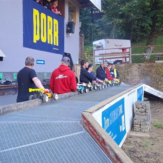 Mehrere Personen beobachten ein Rennen von Miniaturautos von der Seitenlinie aus. Die Autos sind auf einer Rennstrecke aufgereiht, während Zuschauer zusehen. Ein Gebäude mit einem Schild, auf dem 'DORR' steht, ist sichtbar.