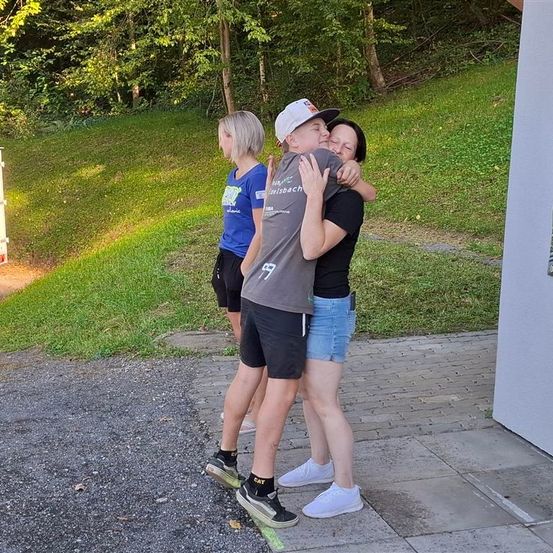 Drei Personen stehen auf einem gepflasterten Weg. Zwei umarmen sich, während eine Frau hinter ihnen steht. Sie sind von einem Grasbereich mit Bäumen umgeben.