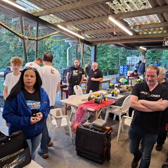 Menschen versammeln sich in einem überdachten Bereich mit Tischen, Werkzeugen und einem Automodell. Ein Mann in einer blauen Jacke steht mit Sonnenbrille auf dem Kopf. Bäume und ein Metalldach sind sichtbar.