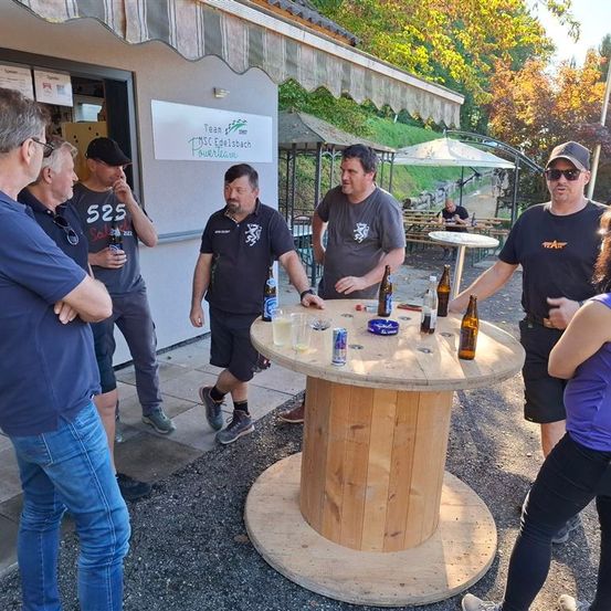 Eine Gruppe von Männern und einer Frau versammelt sich um einen Holztisch mit verschiedenen Getränken und Flaschen. Ein Mann hält eine Bierflasche. Hinter ihnen ein Gebäude mit einem Schild, auf dem Team DSC Edelstach Powerlane steht.