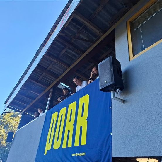 Mehrere Personen beobachten eine Szene von einer Balkonterrasse aus. Ein blauer Banner mit gelben Buchstaben, die 'PORR' lesen, ist an der Wand montiert. Ein Lautsprecher ist neben dem Banner an der Wand befestigt.