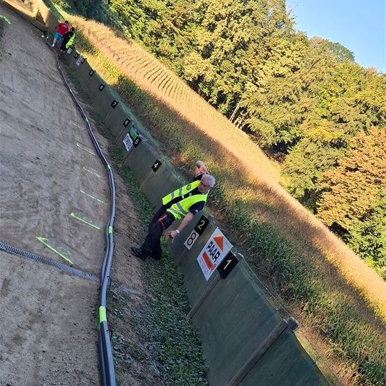 Ein Mann in einer Sicherheitsweste lehnt sich gegen eine Barriere auf einer Erdrennbahn. Zwei Personen gehen in der Nähe. Auf der Barriere sind Zahlen markiert. Im Hintergrund sind Bäume und Gras sichtbar.