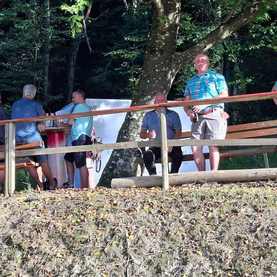 Eine Gruppe von Männern sitzt auf Bänken in einem Waldgebiet, lächelt und schaut zur Kamera. Ein Mann steht in der Nähe eines Baumes.