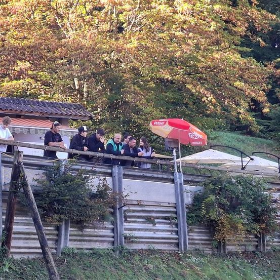 Eine Gruppe von Menschen steht hinter einem Metallzaun in der Nähe eines roten Regenschirms, mit einem Baum mit gelben Blättern im Hintergrund.