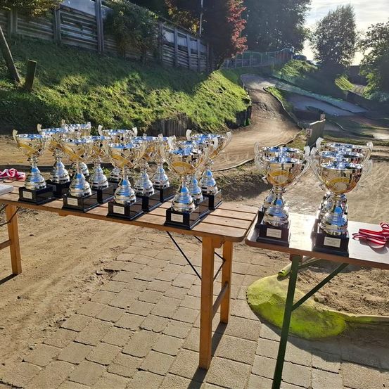 Mehrere silberne Pokale sind auf hölzernen Tischen in einem sandigen Bereich ausgestellt, mit einem schmutzigigen Pfad und einem Zaun im Hintergrund.