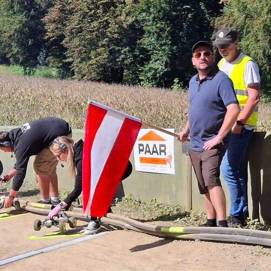 Drei Männer in Freizeitkleidung stehen neben einer Rennstrecke, einer hält eine Flagge. Zwei Personen passen kleine Fahrzeuge auf der Strecke an. Dahinter steht ein Schild mit der Aufschrift 'PAAR'.