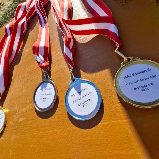 Drei Medaillen und Bänder von der MSC Edelsbach 4 TL-GP Series Süd B-Finale VB 2025 Veranstaltung sind auf einer Holzoberfläche ausgestellt. Jede Medaille hat eine silberne Scheibe mit schwarzen Aufschriften.
