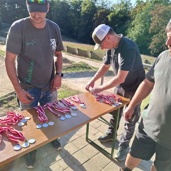 Drei Männer versammeln sich um einen Tisch und betrachten Medaillen mit roten und weißen Bändern. Der Mann auf der linken Seite trägt eine Sonnenbrille und ein graues T-Shirt mit weißer Schrift. Ein anderer Mann in der Mitte trägt eine Kappe und eine Brille, und der Mann auf der rechten Seite trägt ein schwarzes Hemd.