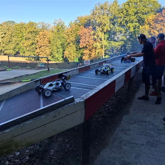 Drei ferngesteuerte Autos sind auf einer schrägen Strecke, wobei ein Mann eines hält. Die Strecke ist von einem Metallzaun umgeben, und es gibt zuschauende Personen. Es gibt Bäume in der Ferne.
