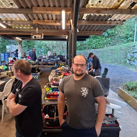 Zwei Männer stehen unter einem Vordach, einer mit verschränkten Armen und der andere mit den Händen in den Taschen. Sie sind von einem unordentlichen Bereich mit Tischen, Stühlen und elektronischer Ausrüstung umgeben. Andere Menschen arbeiten im Hintergrund an Gegenständen. Die Umgebung scheint im Freien zu sein, mit einem Hang und sichtbarer Grünfläche.