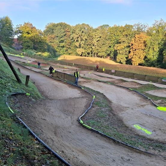 Eine Dirtbike-Strecke mit mehreren Personen im Hintergrund, umgeben von Bäumen und Gras. Die Strecke hat eine Kurve und ist mit gelber Farbe markiert.