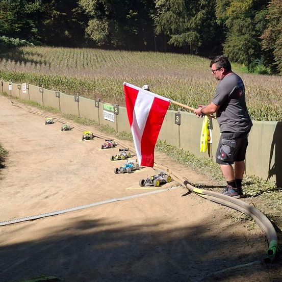 Ein Mann steht neben einer Erdstrecke und hält eine rot-weiße Flagge. Mehrere kleine Autos fahren auf der Strecke, die mit 3 und 5 nummeriert sind. Die Strecke ist von einer Mauer und einem Grasbereich umgeben.