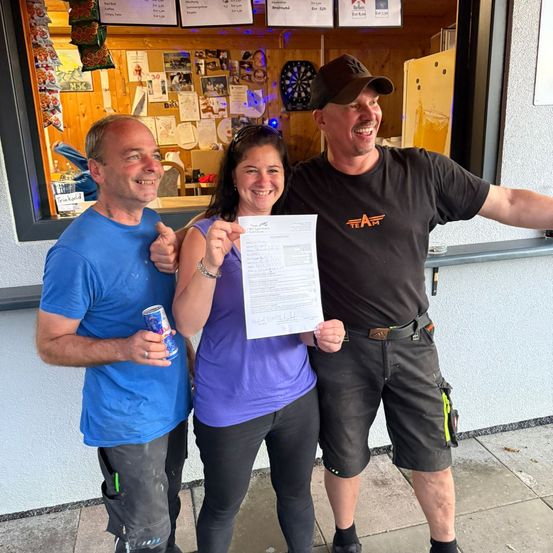 Drei Personen stehen lächelnd vor einem Gebäude. Die Frau hält ein Papier. Der Mann links hält eine Dose. Der Mann rechts trägt einen Hut und hält die Schulter der Frau.