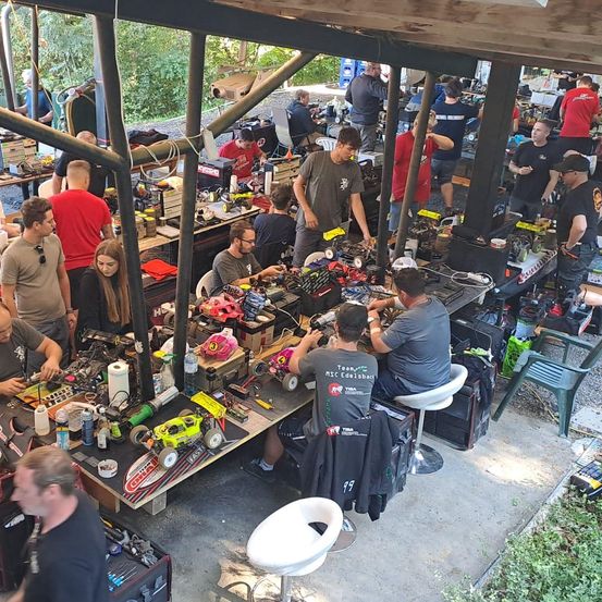 Eine Vogelperspektive auf eine Werkstatt, in der Menschen an Projekten an Tischen arbeiten, umgeben von Werkzeugen und Ausrüstung, unter einem Holzdach mit Bäumen im Hintergrund.