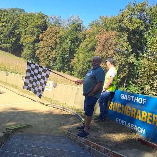 Ein Mann hält eine gemusterte Flagge in der Nähe einer Erdstrecke. Ein weiterer Mann in einer reflektierenden Weste steht hinter ihm. Ein blauer Banner steht für 'Gasthof Buchgraber Fädelsbach.' Bäume säumen den Hintergrund.