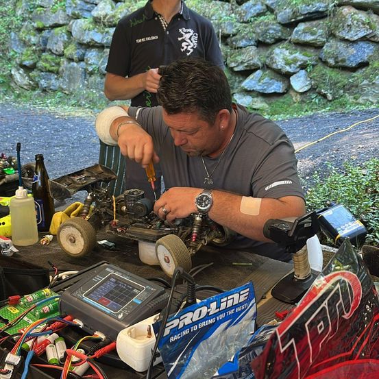 Ein Mann in einem grauen T-Shirt arbeitet mit Werkzeugen und Kabeln an einem ferngesteuerten Auto. Eine Pro-Line Racing Tasche ist in der Nähe. Hinter ihm steht eine weitere Person in einem schwarzen Shirt gegen eine Steinmauer.