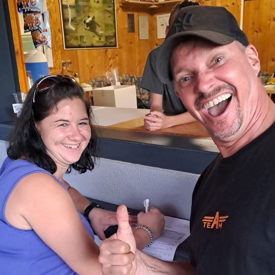 Eine Frau in einem blauen Tanktop und ein Mann in einem schwarzen Shirt mit orangem Text lächeln und geben einen Daumen hoch.