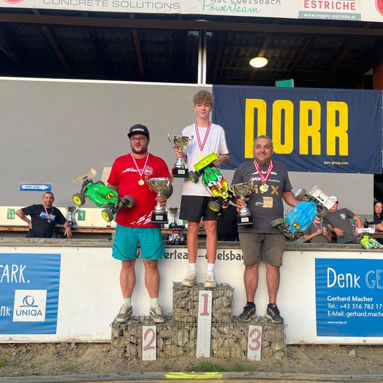 Drei Männer stehen auf einem Podest mit Pokalen in der Hand. Einer hält ein grünes Fahrzeug. Hinter ihnen steht ein Schild mit der Aufschrift 'DORR'. Im Hintergrund beobachtet ein Mann in Schwarz.