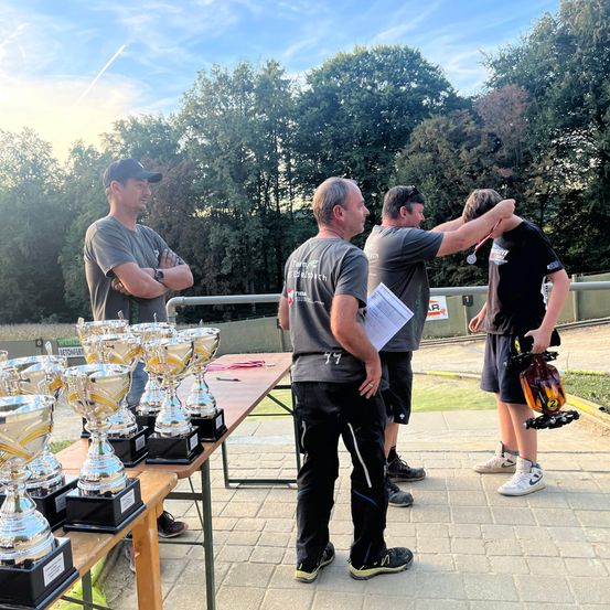 Eine Gruppe von Männern in Freizeitkleidung steht bei einem Tisch mit Trophäen. Sie gratulieren einem jungen Jungen, der eine Medaille um den Hals trägt.