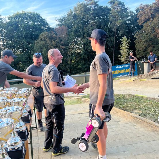 Männer stehen um einen Tisch mit Trophäen und schütteln Hände, ein Junge hält ein Spielzeugauto.