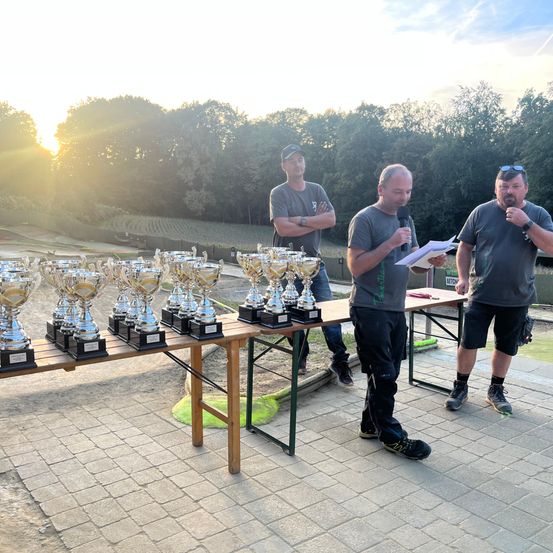 Drei Männer stehen in der Nähe eines Tisches mit vielen Pokalen, wahrscheinlich während einer Siegerehrung. Ein Mann hält ein Mikrofon und liest von einem Papier vor.