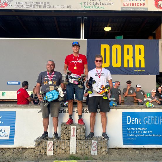 Drei Männer stehen auf einem Podest mit Medaillen und halten ferngesteuerte Autos. Dahinter steht ein Banner mit der Aufschrift 'DORR'. Zuschauer beobachten aus der Ferne.