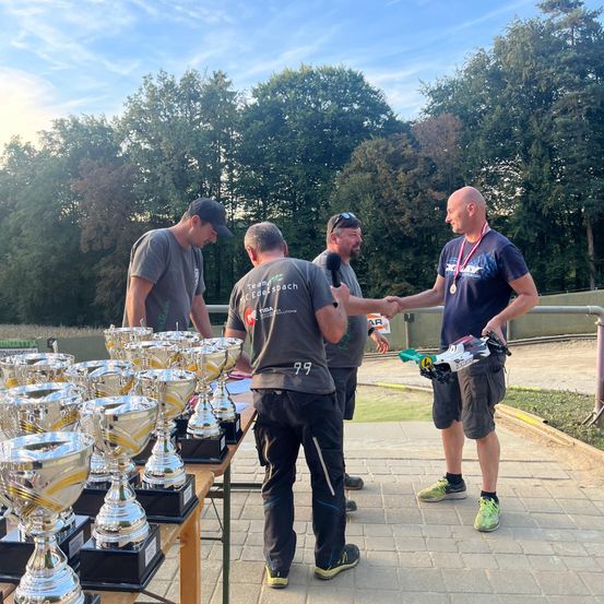 Eine Gruppe von Männern schüttelt sich die Hände vor einem mit Trophäen gefüllten Tisch. Im Hintergrund sind Bäume zu sehen.