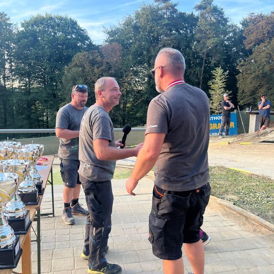 Drei Männer stehen auf einem gepflasterten Weg mit Trophäen. Ein Mann hält ein Mikrofon. Ein anderer trägt eine rot-weiße Namensplakette. Bäume und ein blauer Banner sind im Hintergrund.