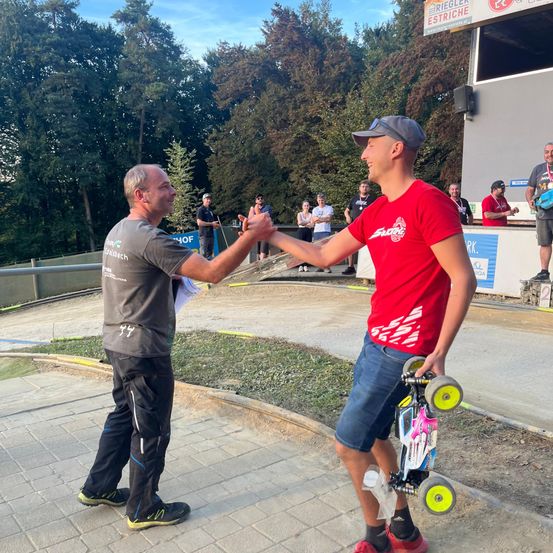 Zwei Männer schütteln sich die Hände bei einer Veranstaltung im Freien. Ein Mann hält ein ferngesteuertes Auto. Zuschauer und Bäume sind im Hintergrund.