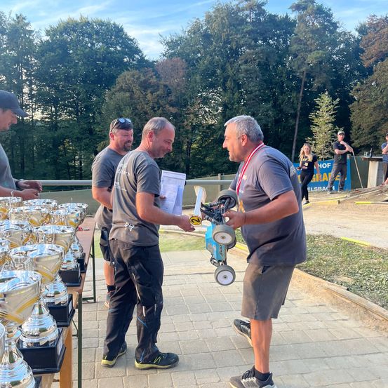 Mehrere Männer stehen um einen Tisch mit Trophäen herum. Ein Mann hält ein kleines Rennauto, während ein anderer ein Stück Papier in der Hand hat. Sie lächeln und scheinen einen Sieg zu feiern.