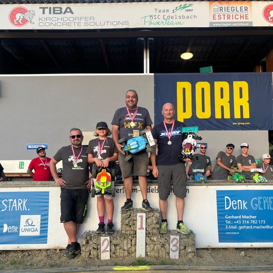 Eine Gruppe von Menschen steht auf einem Podium, einer hält ein Spielzeugauto. Sie tragen Medaillen und halten eine Trophäe. Dahinter sitzen und stehen Menschen, von denen einige ebenfalls Medaillen tragen. Der Bereich hat ein Banner mit dem Wort Dorr.
