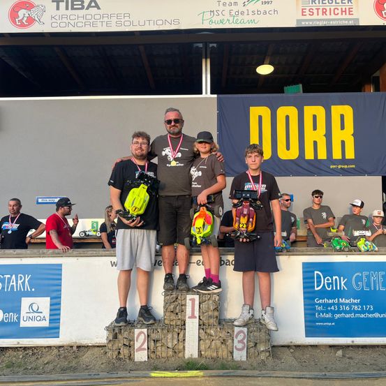 Drei Männer stehen auf einem Podest, halten Pokale und Modelle von Autos. Einer trägt eine Kappe, ein anderer hat eine Sonnenbrille auf, und der dritte hält eine Kamera. Hinter ihnen schaut eine Menge zu, und ein Banner mit dem Wort Dorr ist sichtbar.